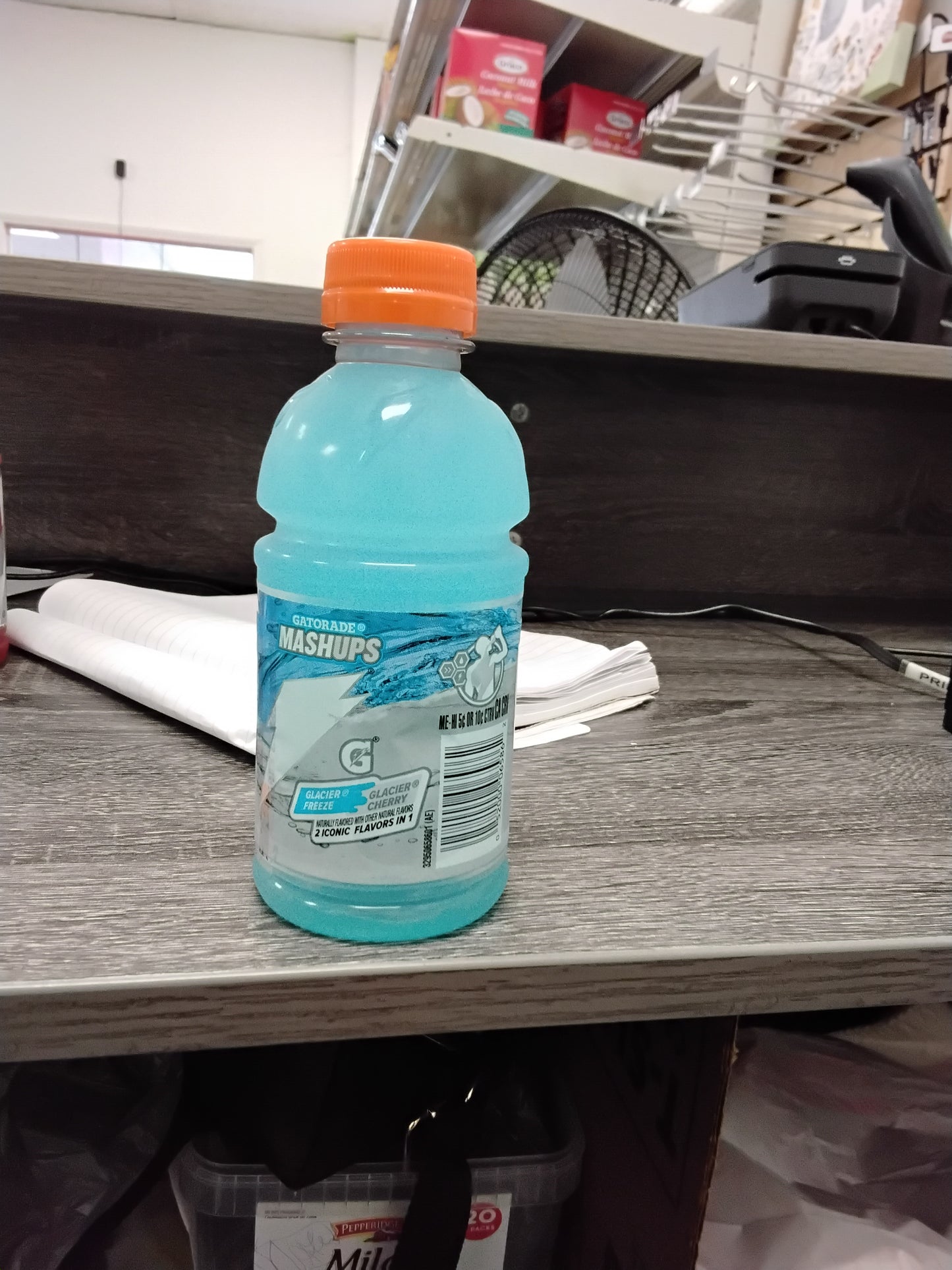 A TOJ EXPRESS Gatorade MASHUPS Glacier Freeze/Glacier Cherry (12 OZ) drink with an orange cap sits on a gray counter, with shelves, papers, and office supplies in the background.