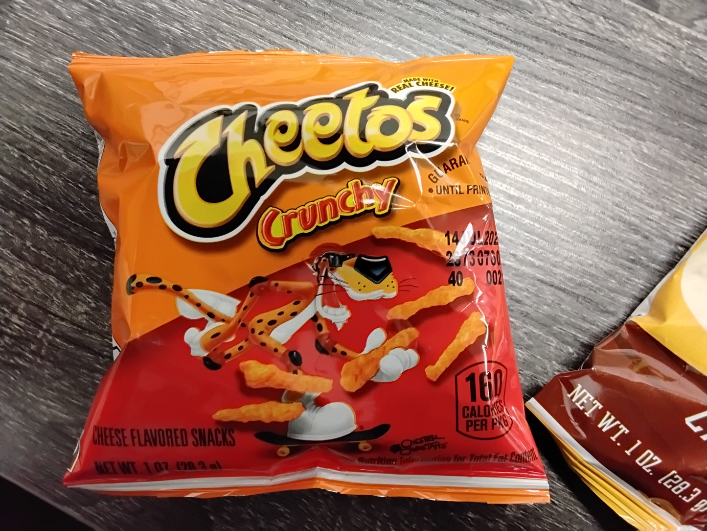 A 1 oz bag of TOJ EXPRESS Cheetos Crunchy Cheese Flavor rests on a gray wooden surface, featuring bold cheesy branding and product info with Chester Cheetah on the orange and red packaging.