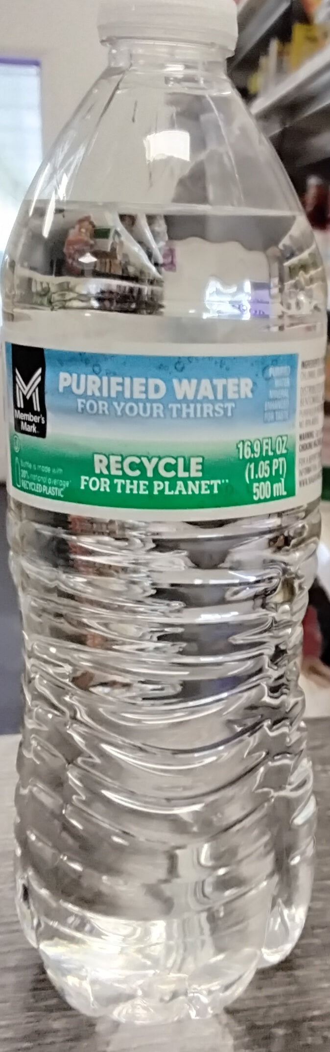A clear plastic bottle of Member's Mark Purified Water 16.9 Oz by TOJ EXPRESS features a blue and green label and offers convenient hydration, shown sitting on a gray indoor surface.