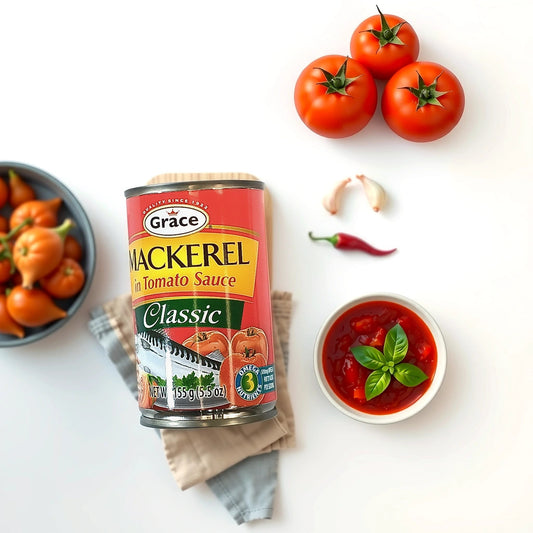 The TOJ EXPRESS Grace Mackerel in Tomato Sauce (1.55 oz) is artfully surrounded by fresh tomatoes, garlic cloves, a red chili, and a bowl of sauce with basil on a white backdrop. A blue bowl of tomatoes and cloth napkins add inspiration for a healthy meal.