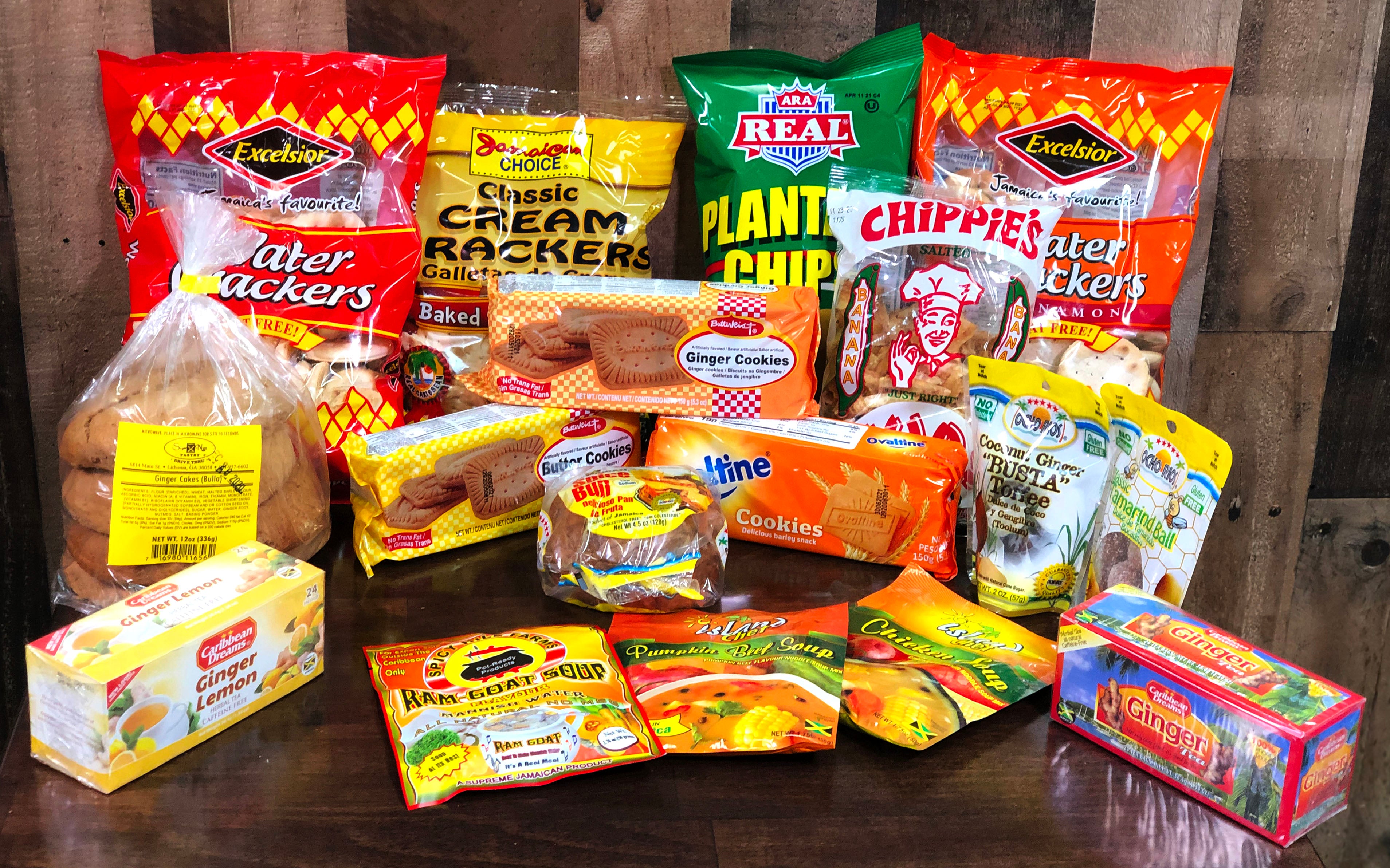 A collection of various snacks and food packages arranged on a wooden surface. The assortment includes crackers, chips, cookies, and seasonings in colorful packaging from different brands.