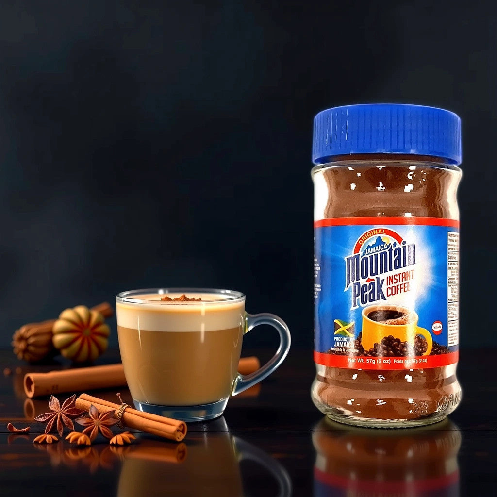 A jar of TOJ EXPRESS Mountain Peak Instant Coffee (2oz) rests beside a clear mug of frothy, rich coffee. Sprinkled with spices, the scene is enhanced by star anise and cinnamon sticks artfully arranged on a glossy black surface.