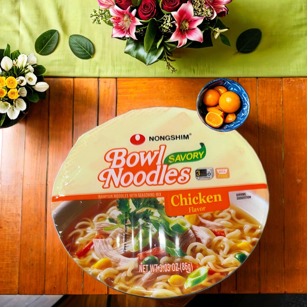 A TOJ EXPRESS NONGSHIM BOWL NOODLES-CHICKEN cup sits on a wooden table with flowers, kumquats, and a green runner—ideal for a quick meal or tasty chicken-flavored soup.