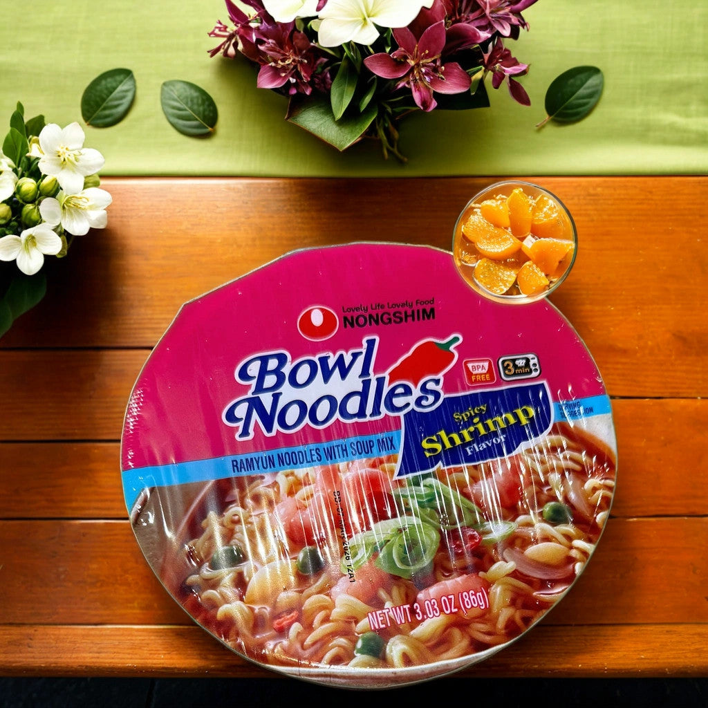 A bowl of TOJ EXPRESS NONGSHIM NOODLES-SHRIMP sits on a wooden table, accompanied by a glass of mandarin orange slices with floral decorations in the background.
