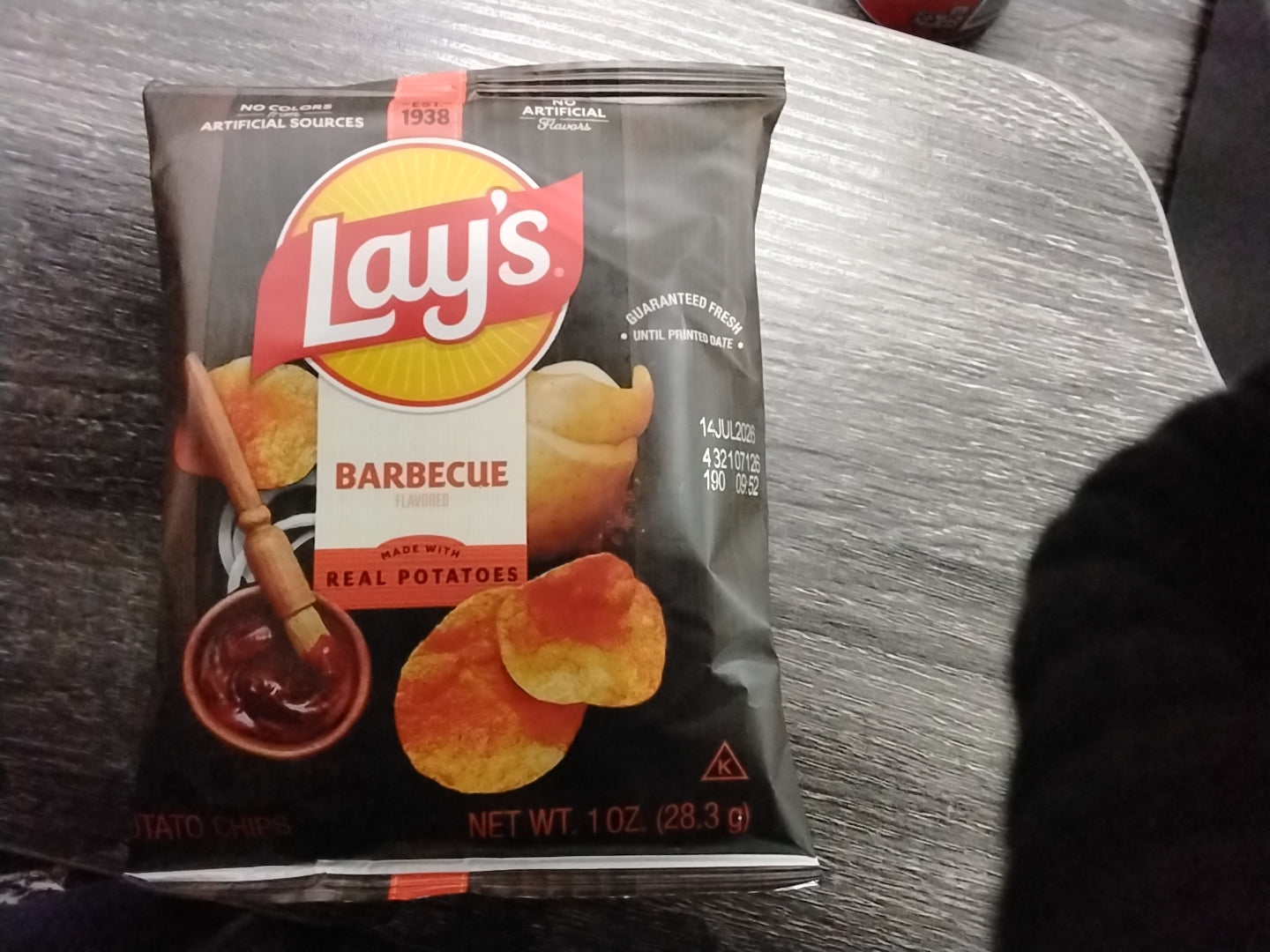A 1 oz bag of TOJ EXPRESS Lays BBQ Chips rests on a table in its black packaging, displaying chips and barbecue sauce. A curious cat is partly visible at the lower right edge of the image.