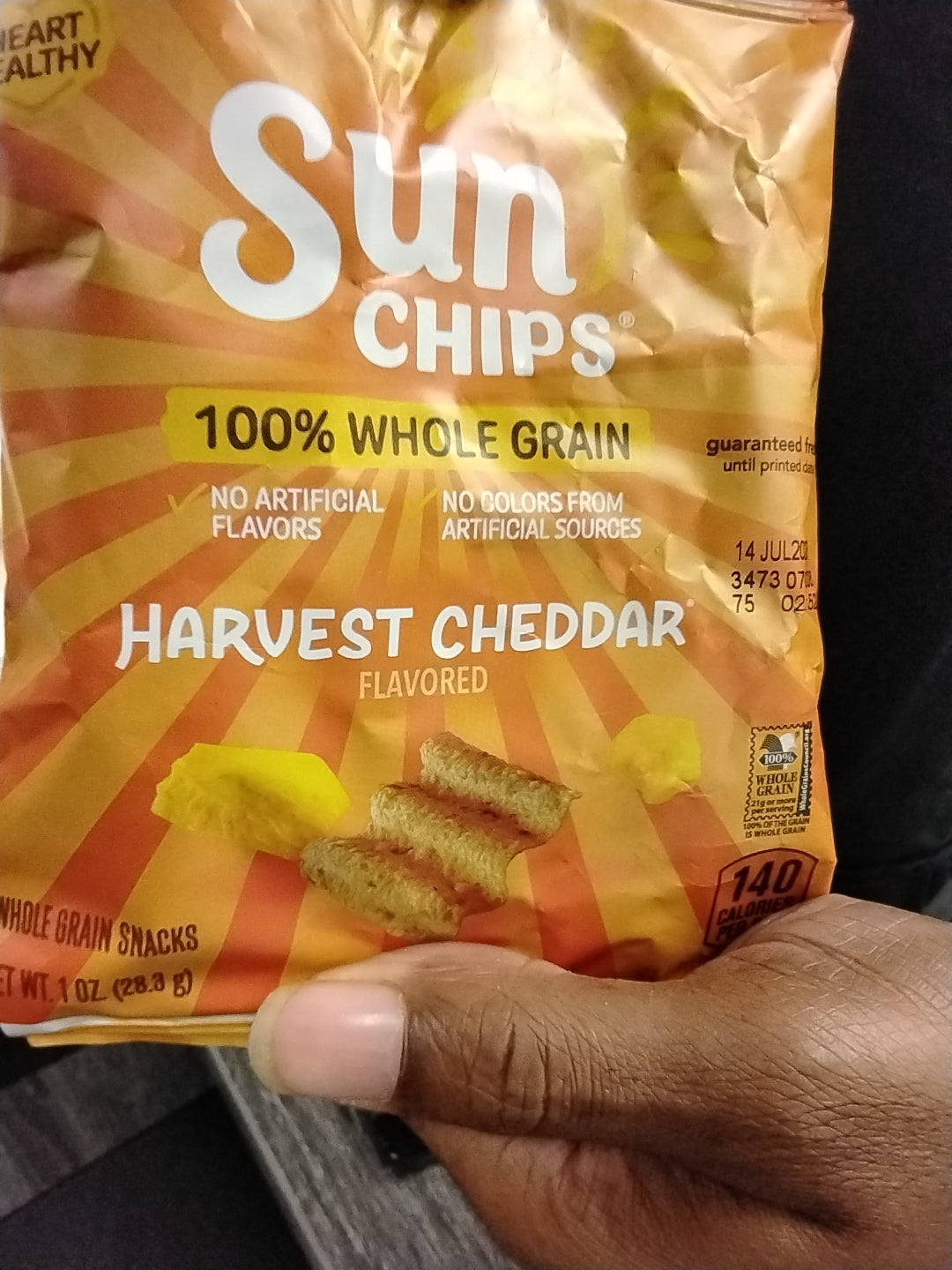 A hand holds a bag of TOJ EXPRESS Sun Chips Harvest Cheddar. The orange and yellow packaging shows the chips and nutrition info, presenting a tasty, whole grain snack option.