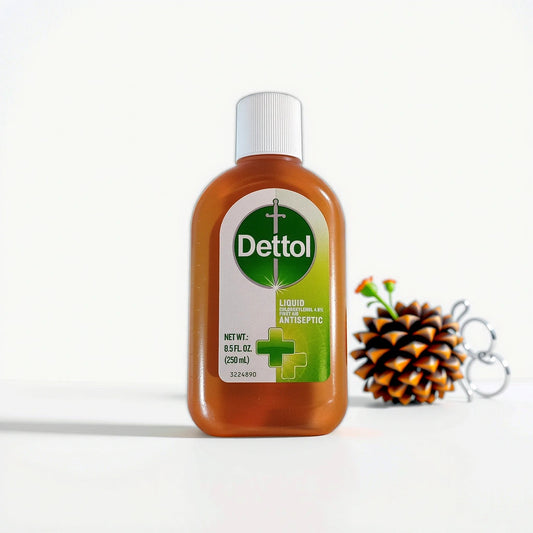A brown bottle of TOJ EXPRESS Dettol Liquid Antiseptic, capped in white, rests on a white surface. The label shows the brand name, volume, and green cross motif, assuring a 99.9% germ kill rate. A pine cone and glasses are elegantly arranged behind it.