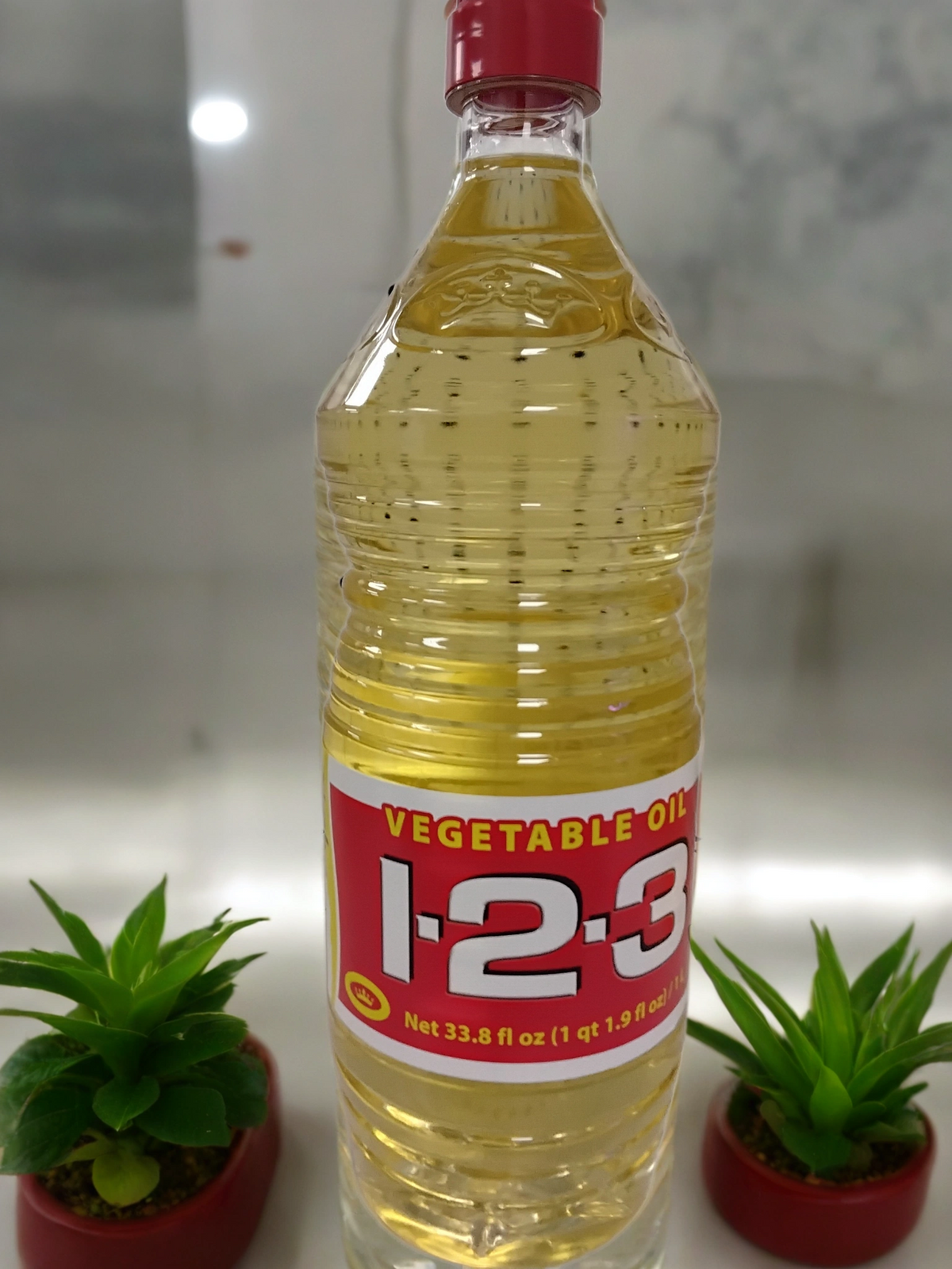 A clear plastic bottle of 1-2-3 vegetable oil with a red label stands between two small green succulent plants in red pots on a white surface.
