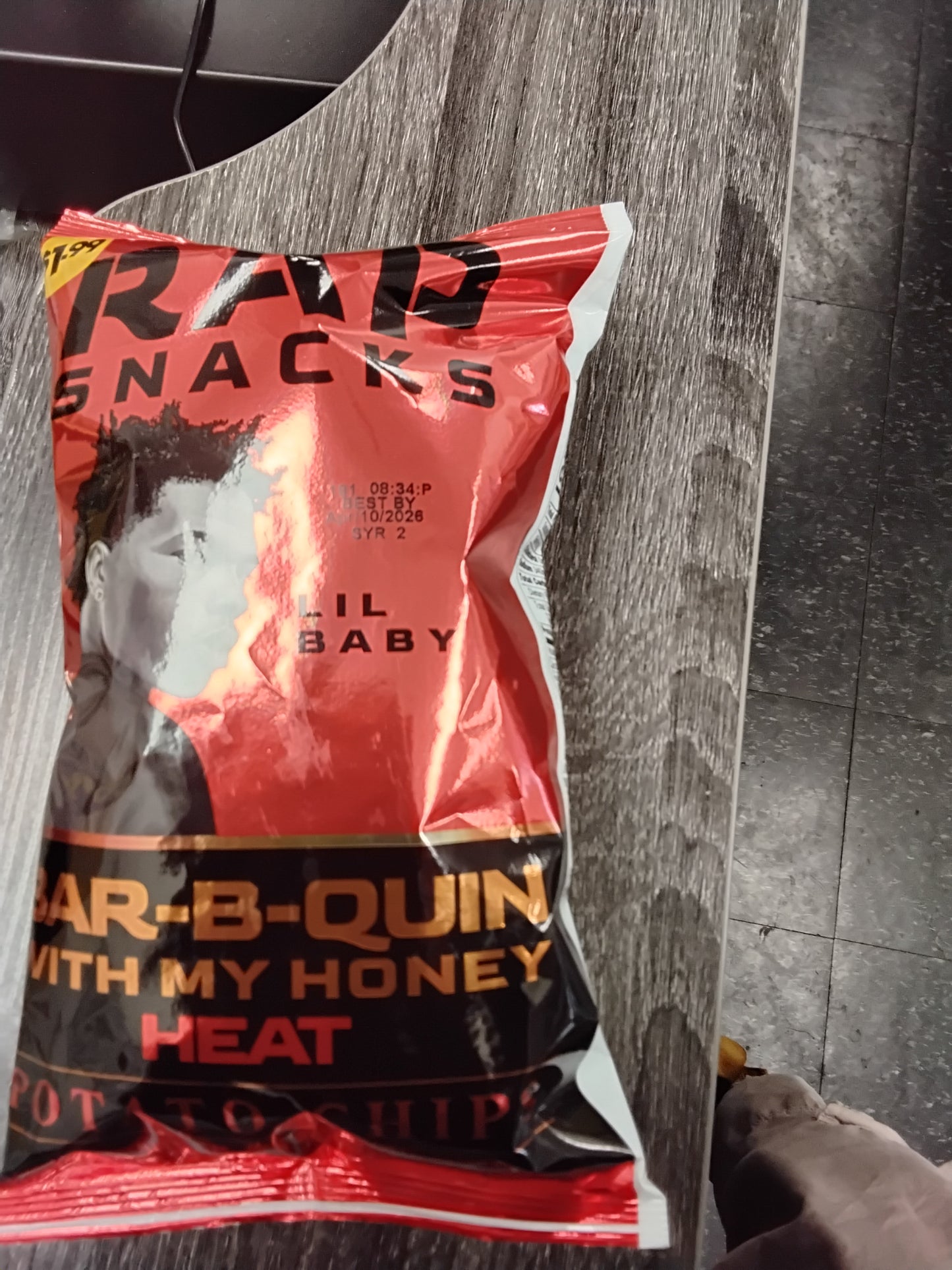 A bag of TOJ EXPRESS Rap Snacks Lil Baby Spicy BBQ potato chips, in red, black, and gold packaging, is placed on a gray wooden surface.