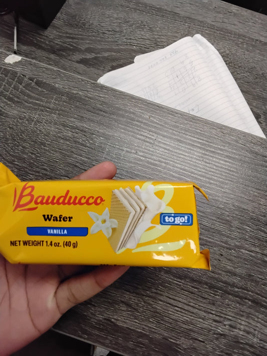 A hand holds a yellow pack of TOJ EXPRESS Bauducco Wafer - Vanilla, featuring vanilla flowers and wafer images on the wrapper. In the background, a notepad with handwritten notes rests on a dark wooden surface.