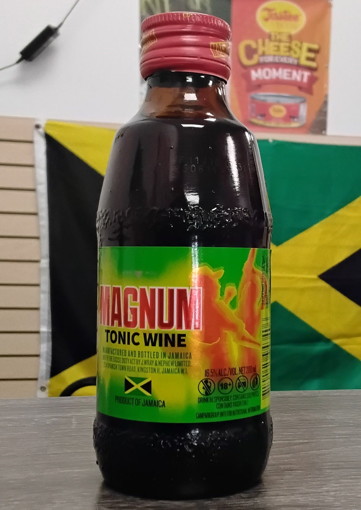 A TOJ EXPRESS Magnum Tonic Wine bottle with a red cap and vibrant yellow, green, and red label sits on a table. Behind it is a Jamaican flag and a Cheese Moment poster on the wall.