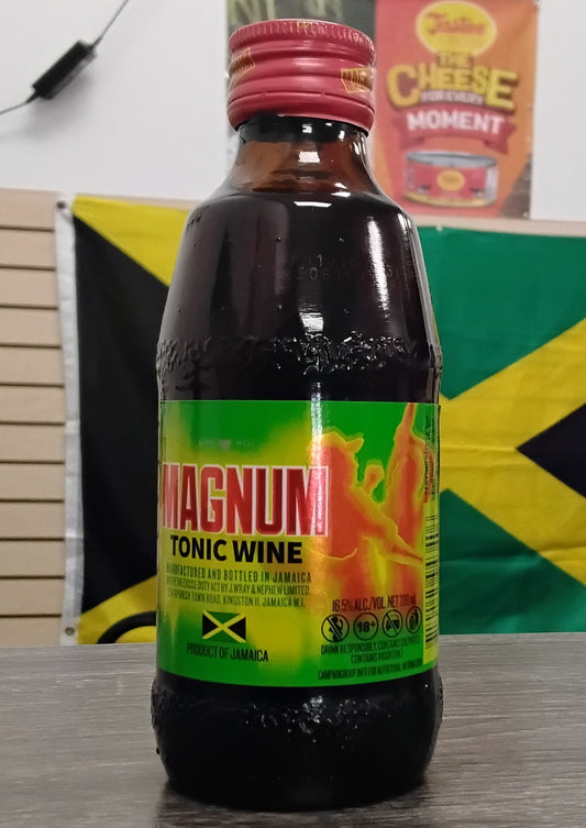 A TOJ EXPRESS Magnum Tonic Wine bottle with a red cap and vibrant yellow, green, and red label sits on a table. Behind it is a Jamaican flag and a Cheese Moment poster on the wall.