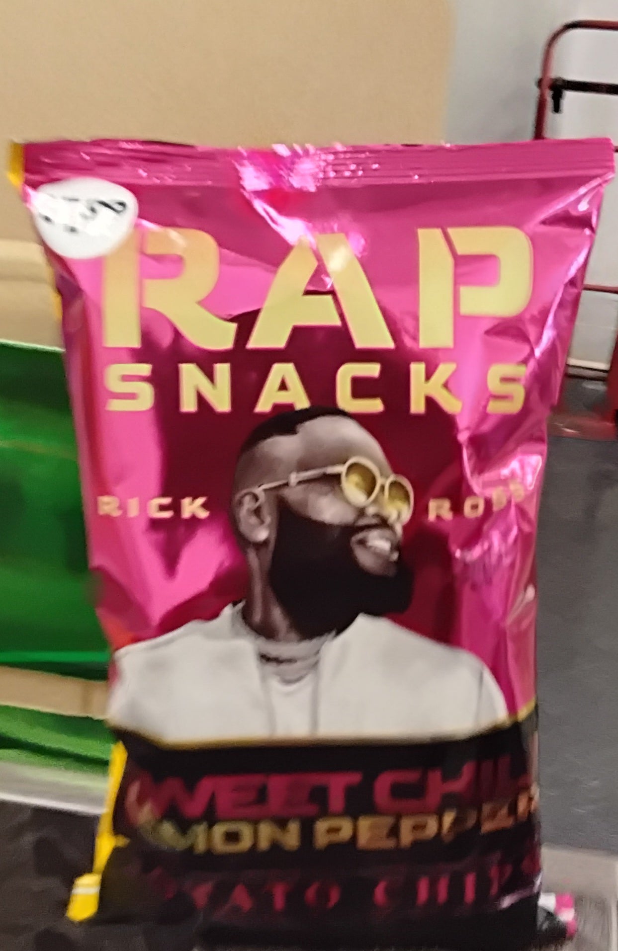 A shiny pink bag of TOJ EXPRESS Rap Snacks Rick Ross Sweet Chili Lemon Pepper Potato Chips featuring an illustrated man with a beard and round glasses.