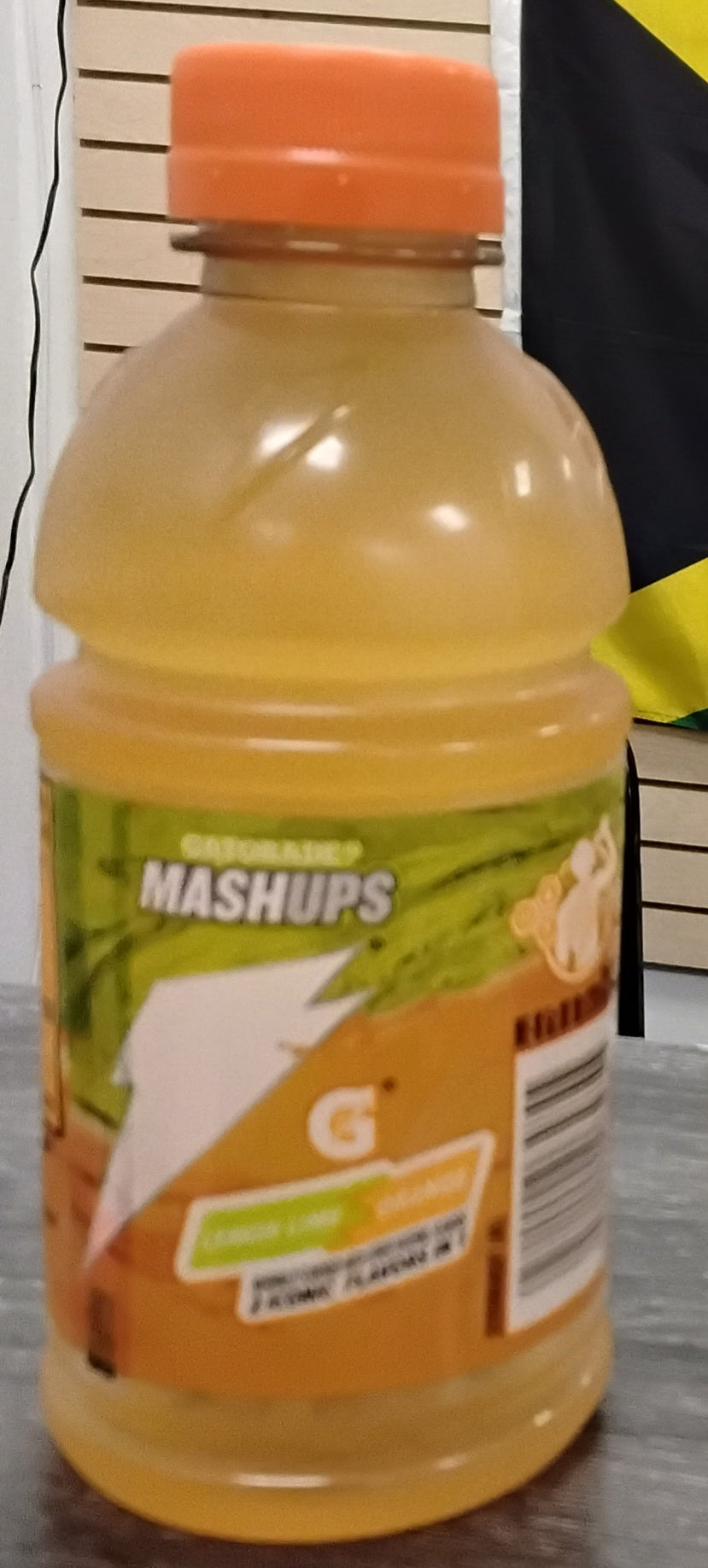 A 12 oz bottle of TOJ EXPRESS Gatorade Mashup Edition LemonLime/Orange with an orange cap sits on a table. The label is orange and green, with a partial Jamaican flag visible in the background.