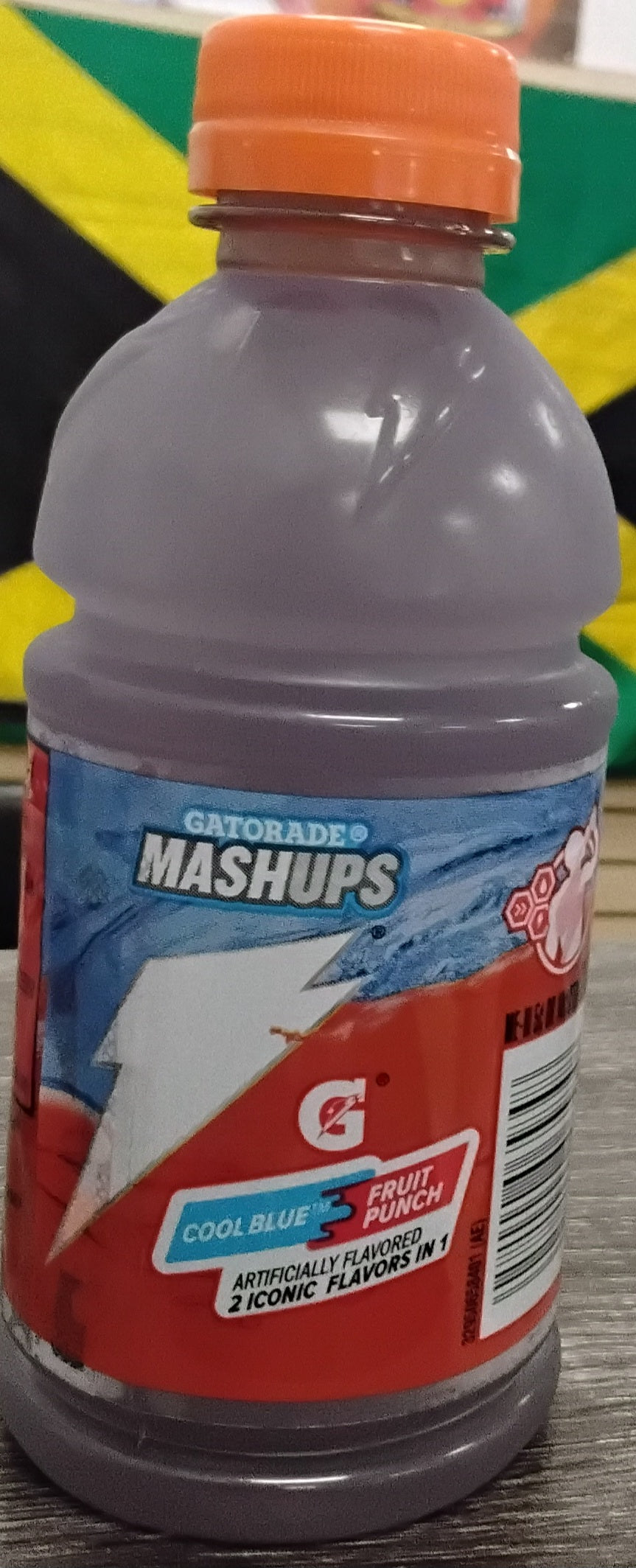A TOJ EXPRESS Gatorade Mashups Edition Cool Blue/Fruit Punch (12 oz) bottle with a purple body and orange cap sits on a table, with a Jamaican flag blurred in the background for a splash of vibrant flair.