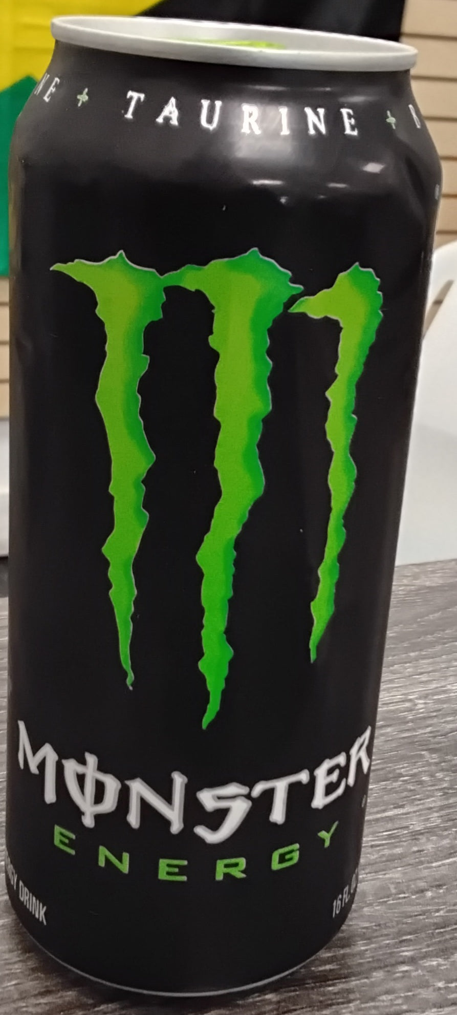 A 16 oz can of TOJ EXPRESS Monster Energy Drink sits on a table. The black can features a large green claw mark M logo and white Monster Energy text, with "Taurine" visible at the top.