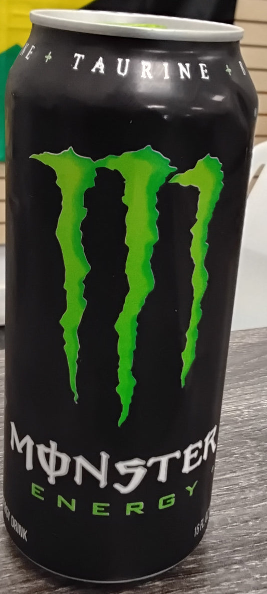 A 16 oz can of TOJ EXPRESS Monster Energy Drink sits on a table. The black can features a large green claw mark M logo and white Monster Energy text, with "Taurine" visible at the top.
