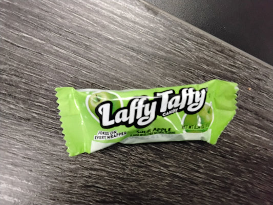 A wrapped piece of TOJ EXPRESS Laffy Taffy (apple) candy, sweet and tart, rests on a dark wood-textured surface. Its chewy green wrapper displays the Laffy Taffy logo in white and black lettering.