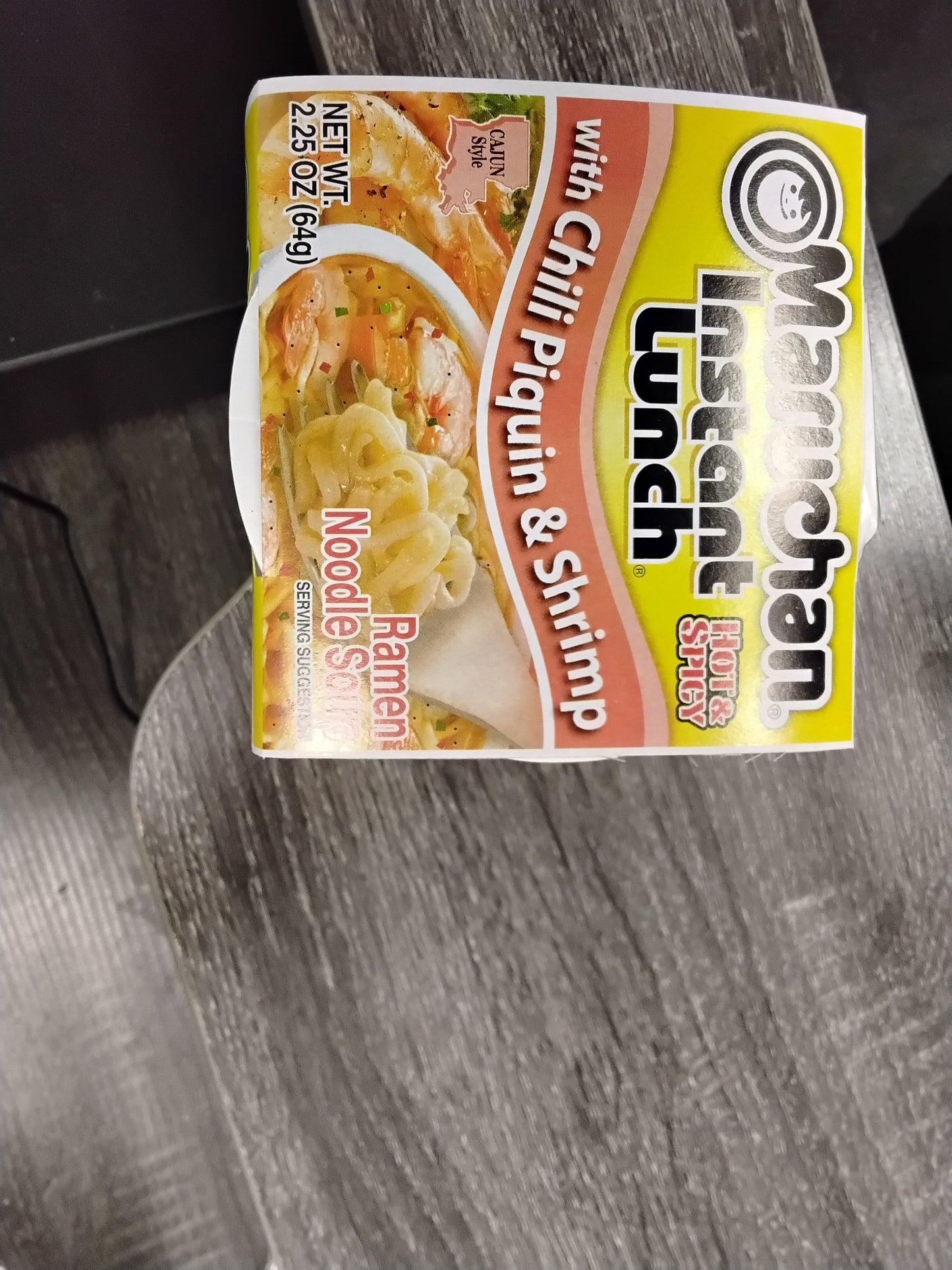 A TOJ EXPRESS Maruchan Instant Cup Noodle Soup Chili w Piquin and Shrimp Flavor sits on a gray wooden surface, in yellow packaging with images of noodles and shrimp, weighing 2.25 oz (64g).
