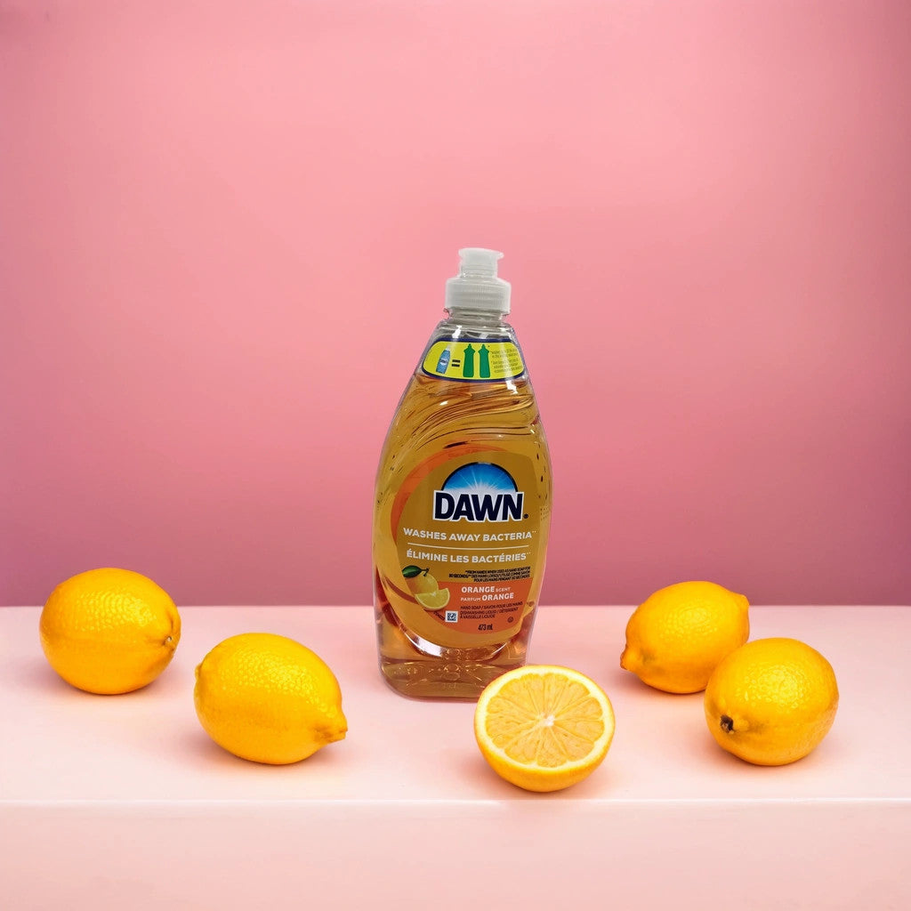A bottle of TOJ EXPRESS Dawn Dishwashing Liquid sits on a pink surface with four whole lemons and one lemon half, set against a pink background, ensuring sparkling clean dishes every wash.