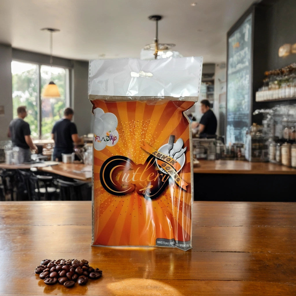A package of TOJ EXPRESS Fundap Cutlery sits on a wooden café table, coffee beans scattered around. The softly blurred background reveals two people and shelves with jars and bottles behind the picnic utensils.