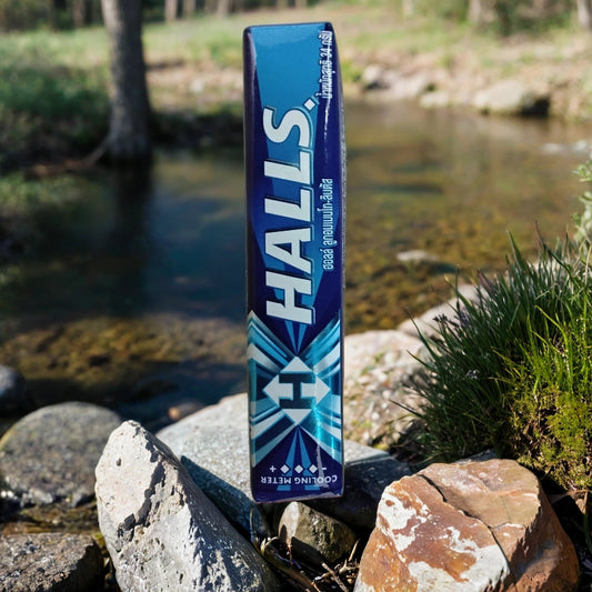 A box of TOJ EXPRESS Halls cough Drops-MENTHOLYPTOS stands upright on rocks by a clear stream, with green grass and trees in the background on a sunny day.