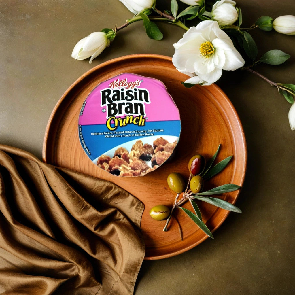 A 2.8 oz bowl of TOJ EXPRESS Kellogg’s Raisin Bran Crunch, with sweet raisins and crunchy bran flakes, is placed on a wooden plate beside a brown cloth, olives on a branch, and white flowers on a green surface.