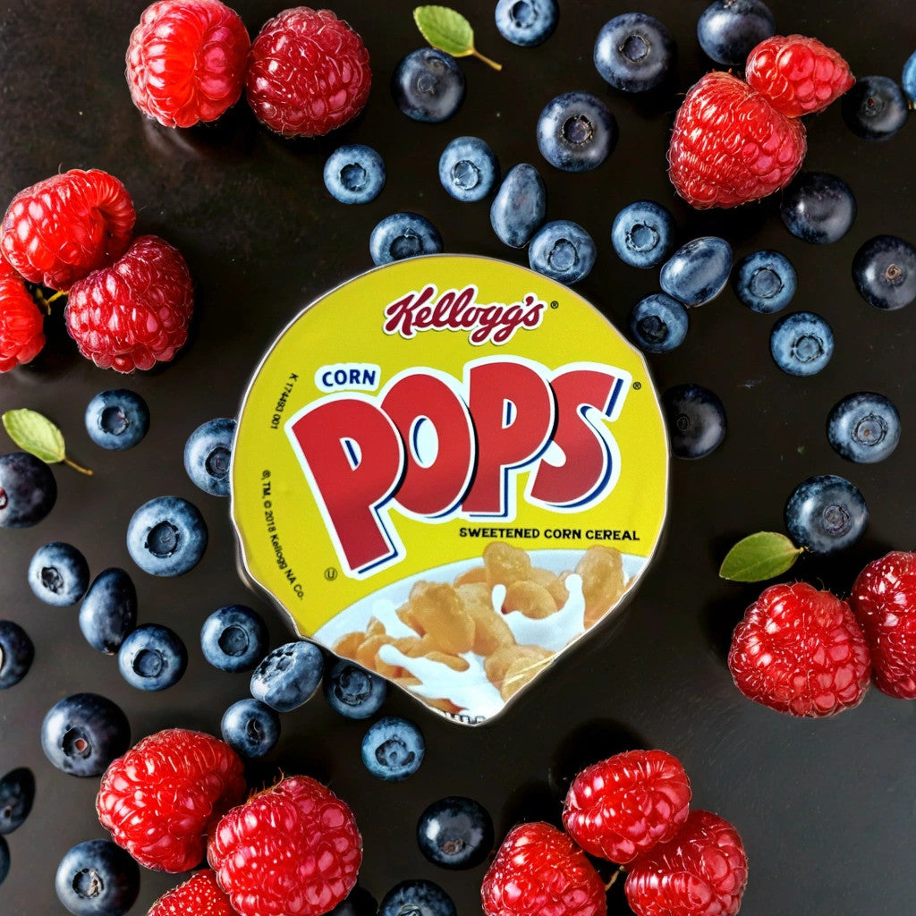 A bowl of TOJ EXPRESS Kellogg's Corn Pops (1.5 oz) breakfast cereal sits on a black surface, surrounded by fresh raspberries, blueberries, and mint leaves.