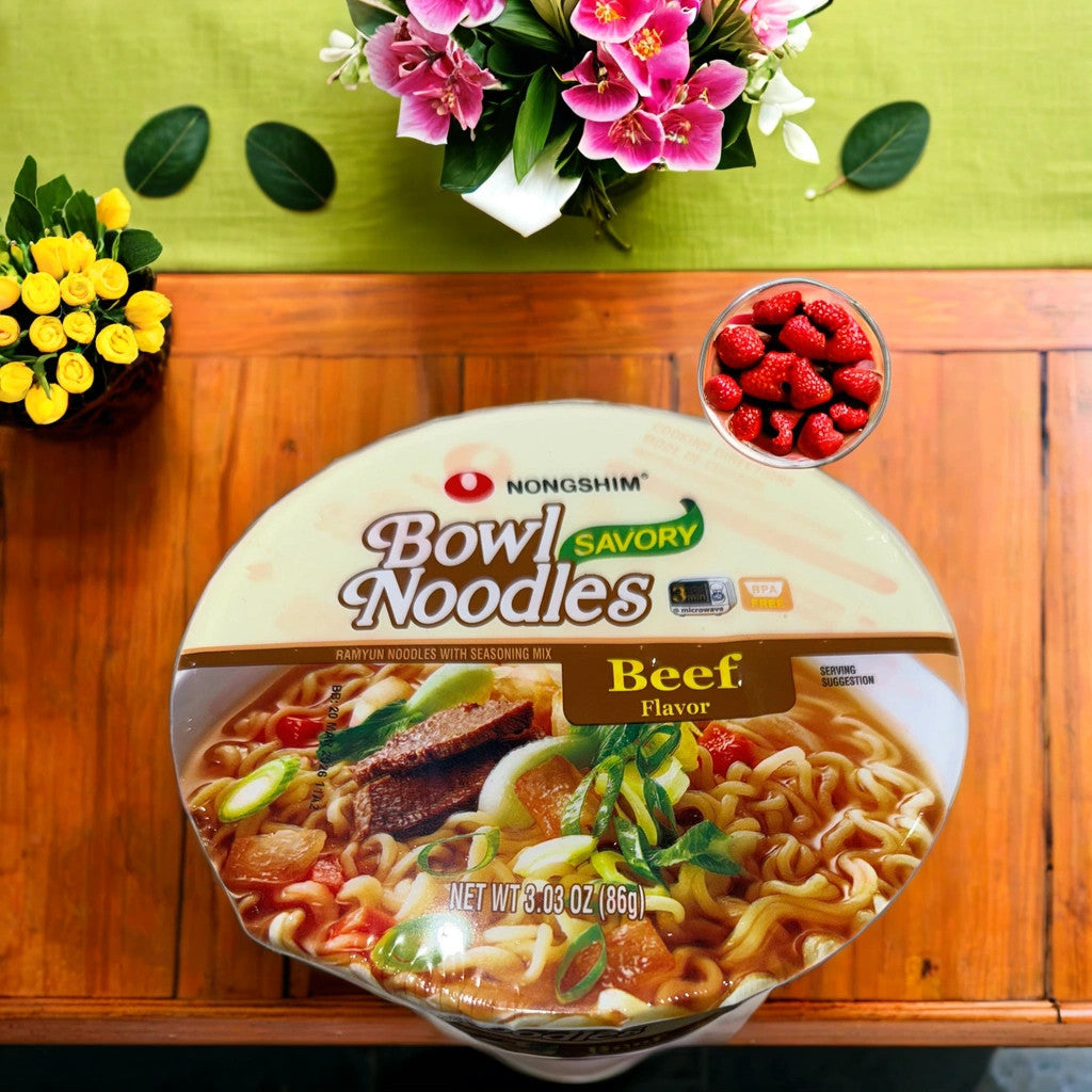 A TOJ EXPRESS NONGSHIM BOWL NOODLES-BEEF sits on a wooden table next to fresh raspberries, yellow roses, and a vase of pink flowers with green leaves, highlighting this tasty instant noodle bowl.