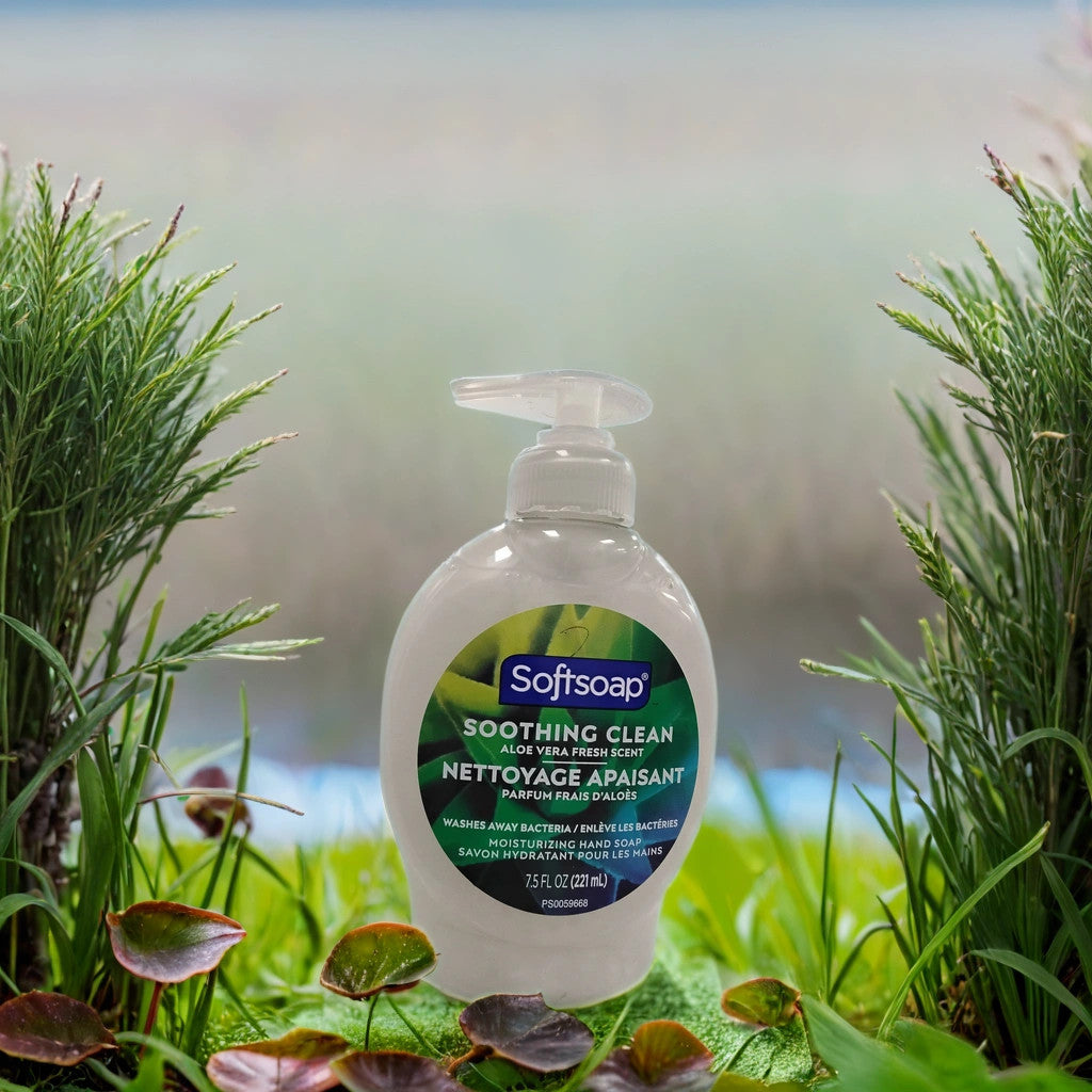 A bottle of TOJ EXPRESS Softsoap (Aloe Vera Fresh Scent) sits outdoors on green leaves, surrounded by grass and plants, with a blurred natural background.