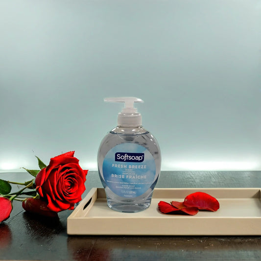 A bottle of TOJ EXPRESS Softsoap-Freshbreeze hand soap sits on a tray beside a red rose and scattered petals, adding a fresh scent to the light blue background.