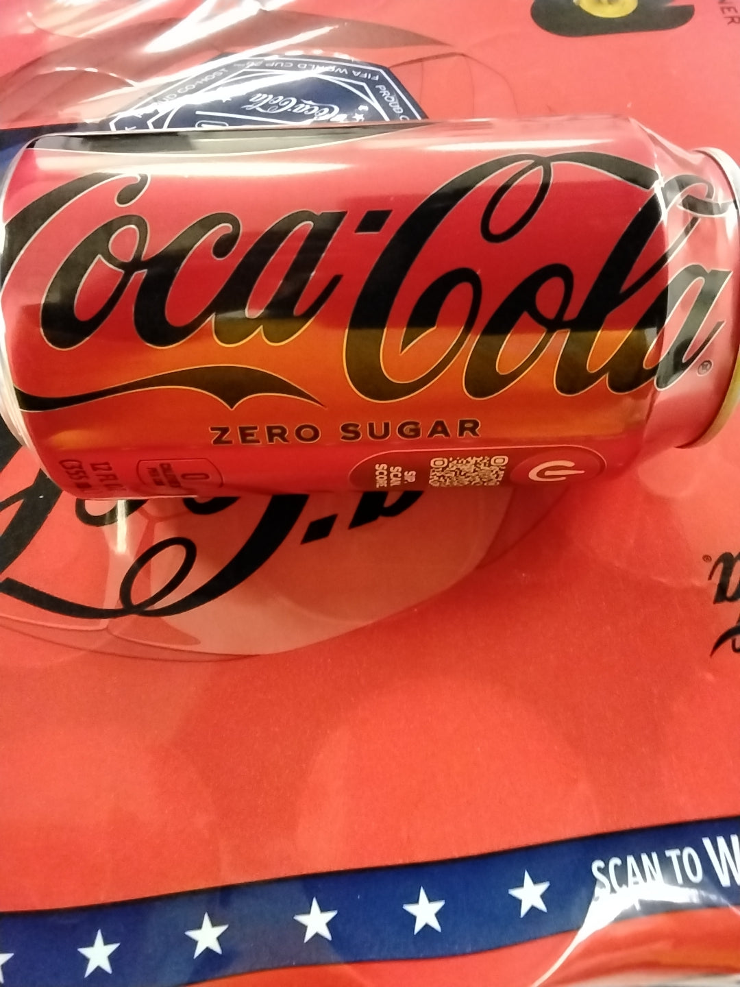A close-up of a TOJ EXPRESS Coca Cola Zero Sugar can, a zero-calorie cola, lying on its side on a red surface with part of a dark blue and white star pattern visible at the bottom.