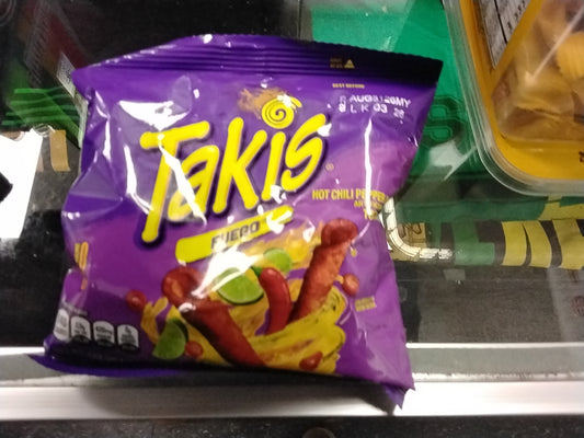 A TOJ EXPRESS Takis - Fuego 1 oz bag sits on a glass counter, displaying its spicy hot chili pepper and lime rolled tortilla chips with fiery images of the chips, limes, and chili peppers on the packaging.