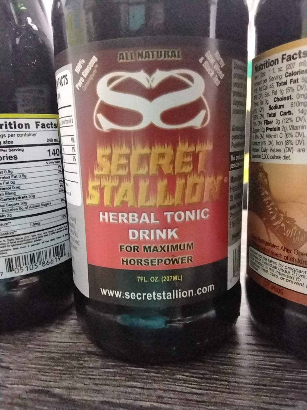 A bottle of Secret Stallion by TOJ EXPRESS, featuring a red and silver design, rests on a dark surface. The performance tonic promises vitality, and nutritional facts are visible on nearby bottles.