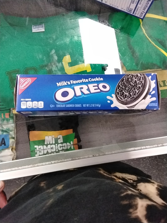 A 5.2 oz package of Nabisco Oreo Chocolate Sandwich Cookies by TOJ EXPRESS is on a glass counter, with the reflection of a green shirt and various items visible beneath the glass.