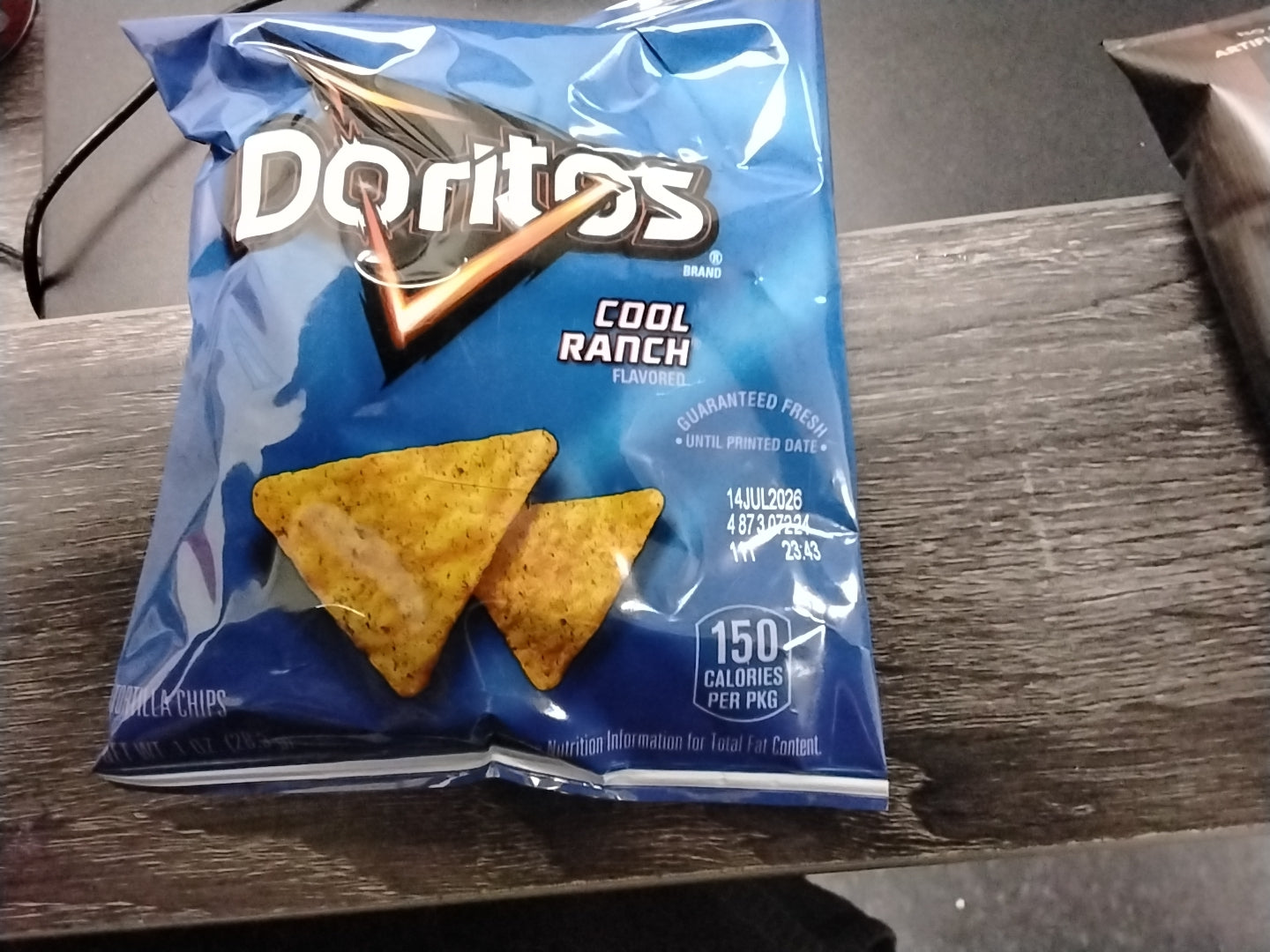 A blue TOJ EXPRESS Doritos Cool Ranch Flavor 1 oz bag rests on a wooden surface, showing two triangular chips and nutrition info with 150 calories per package on the front.
