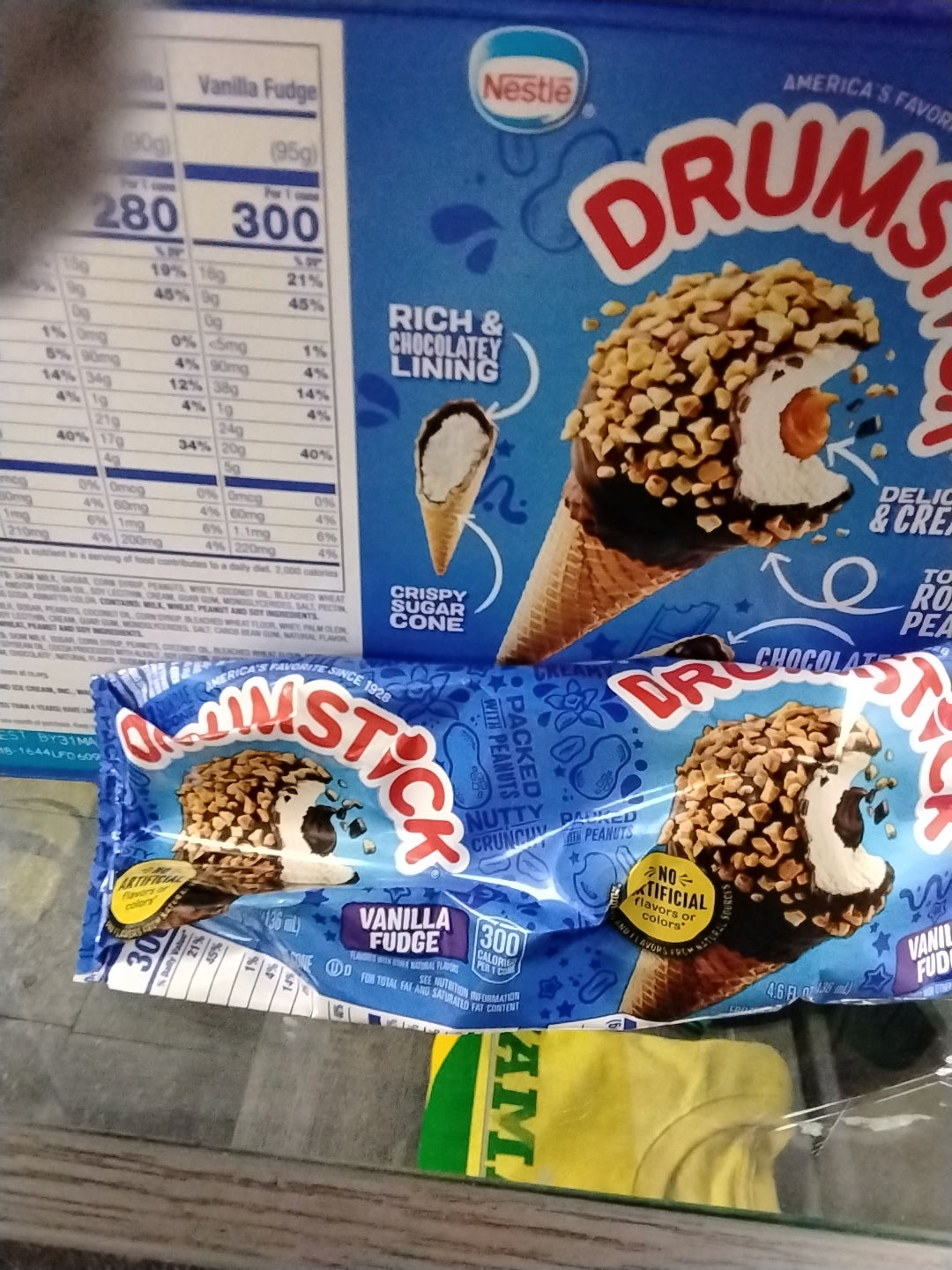 A TOJ EXPRESS DrumStick Ice Cream Vanilla Fudge box featuring a cone image sits on a store shelf next to two blue-wrapped DrumStick cones. Nutrition facts are visible on the box.