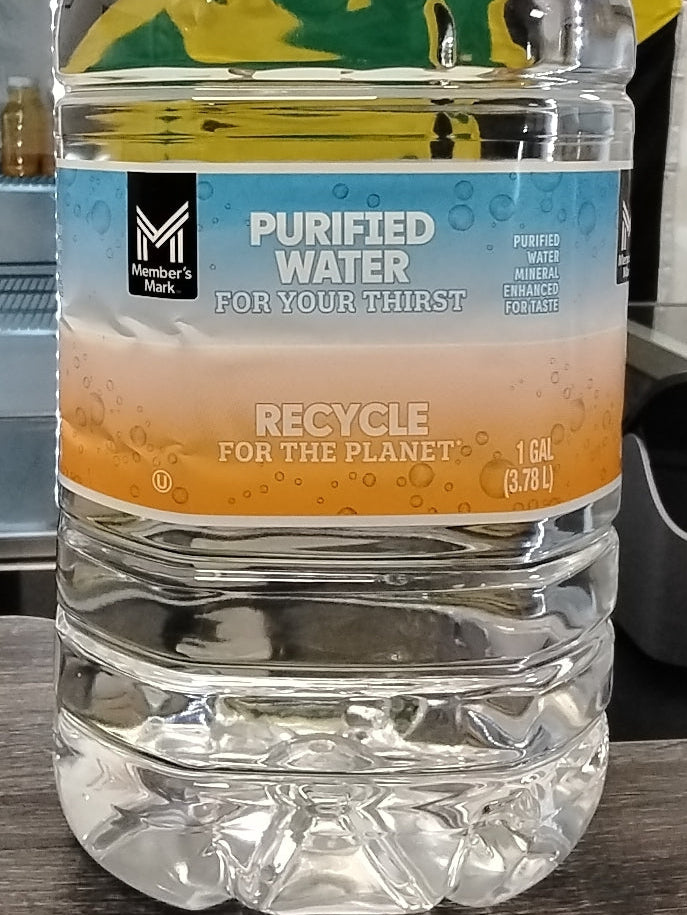A 1-gallon Member's Mark Purified Water bottle by TOJ EXPRESS, designed for recycling and daily hydration, rests on a dark surface.