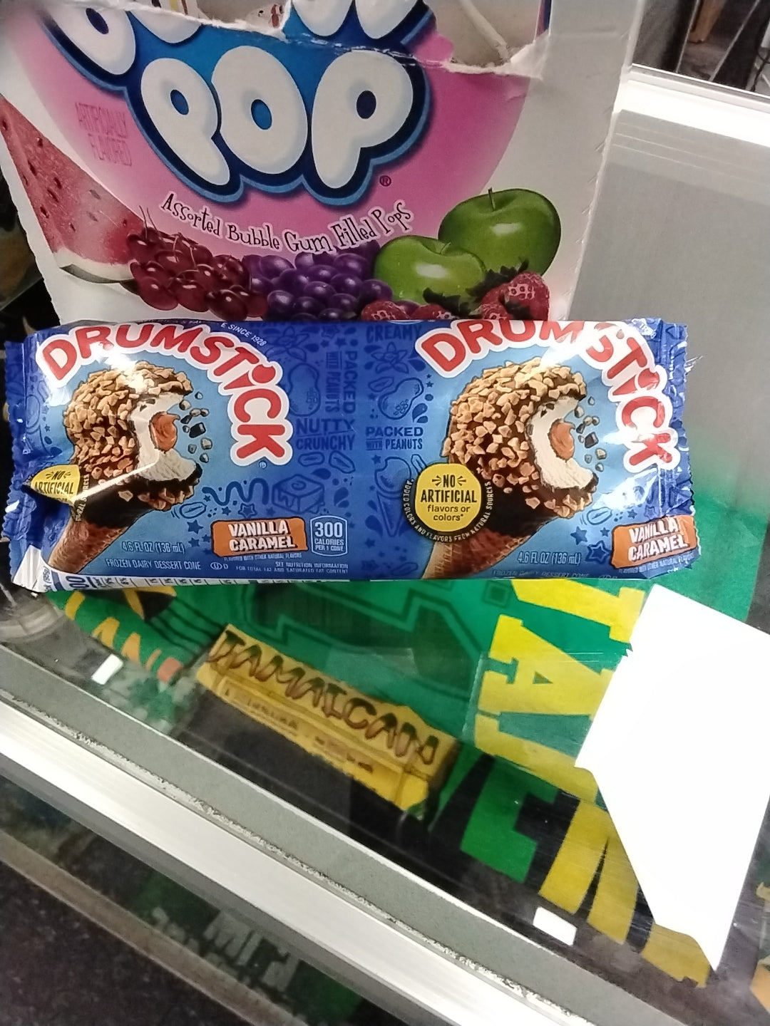 A TOJ EXPRESS DrumStick Ice Cream Vanilla Caramel cone, topped with chocolate, caramel, and nuts, sits on a glass counter in its blue wrapper. In the background, a Dubble Bubble gum box is partially visible.