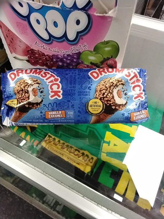 A TOJ EXPRESS DrumStick Ice Cream Vanilla Caramel cone, topped with chocolate, caramel, and nuts, sits on a glass counter in its blue wrapper. In the background, a Dubble Bubble gum box is partially visible.