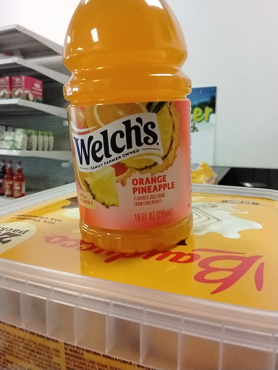 A bottle of TOJ EXPRESS Welches Orange Pineapple Drink rests on a tub of Breyers ice cream in a grocery store aisle lined with shelves of assorted products.