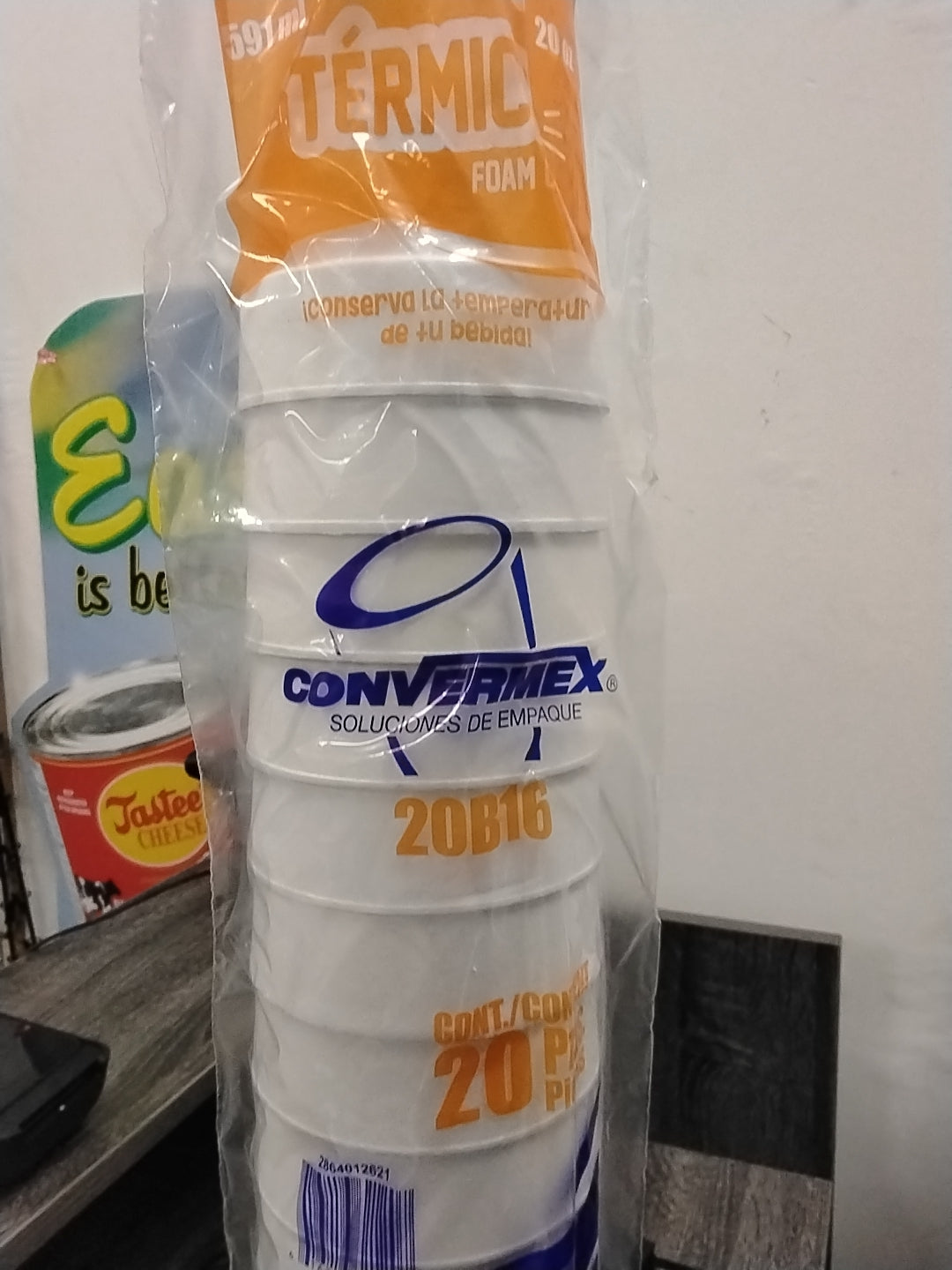 A 20-count pack of TOJ EXPRESS Vaso Termico Foam Cups (20 oz) with thermal insulation, wrapped in plastic. Blue and orange text appears on the cups, along with the "TOJ EXPRESS" brand name. A colorful sign and countertop appear in the background.