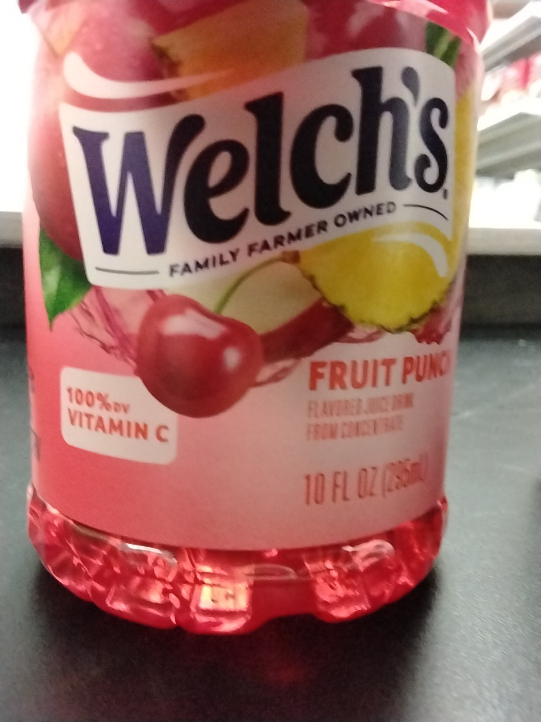 A close-up of a TOJ EXPRESS Welches Fruit Punch Drink label showcases cherries, an apple slice, and highlights 100% DV Vitamin C. The vibrant red label features a refreshing drink rich in essential vitamins and fruit punch flavors.